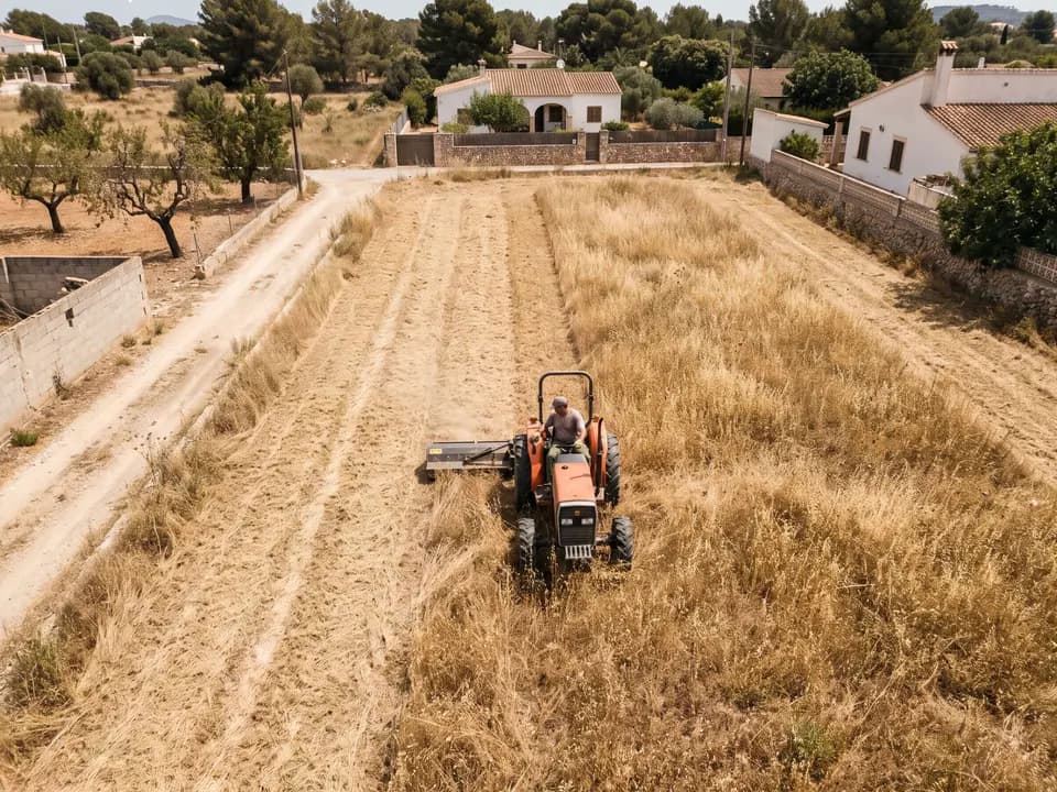 Tractor pequeño con desbrozadora de martillos cortando una parcela llana de hierba seca, vista desde arriba